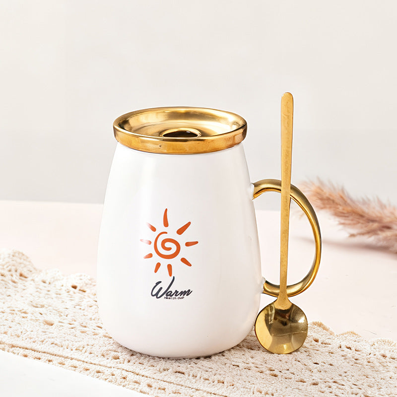 White mug with gold lid and handle, featuring a sun design and 'Warm' text, on a textured surface.