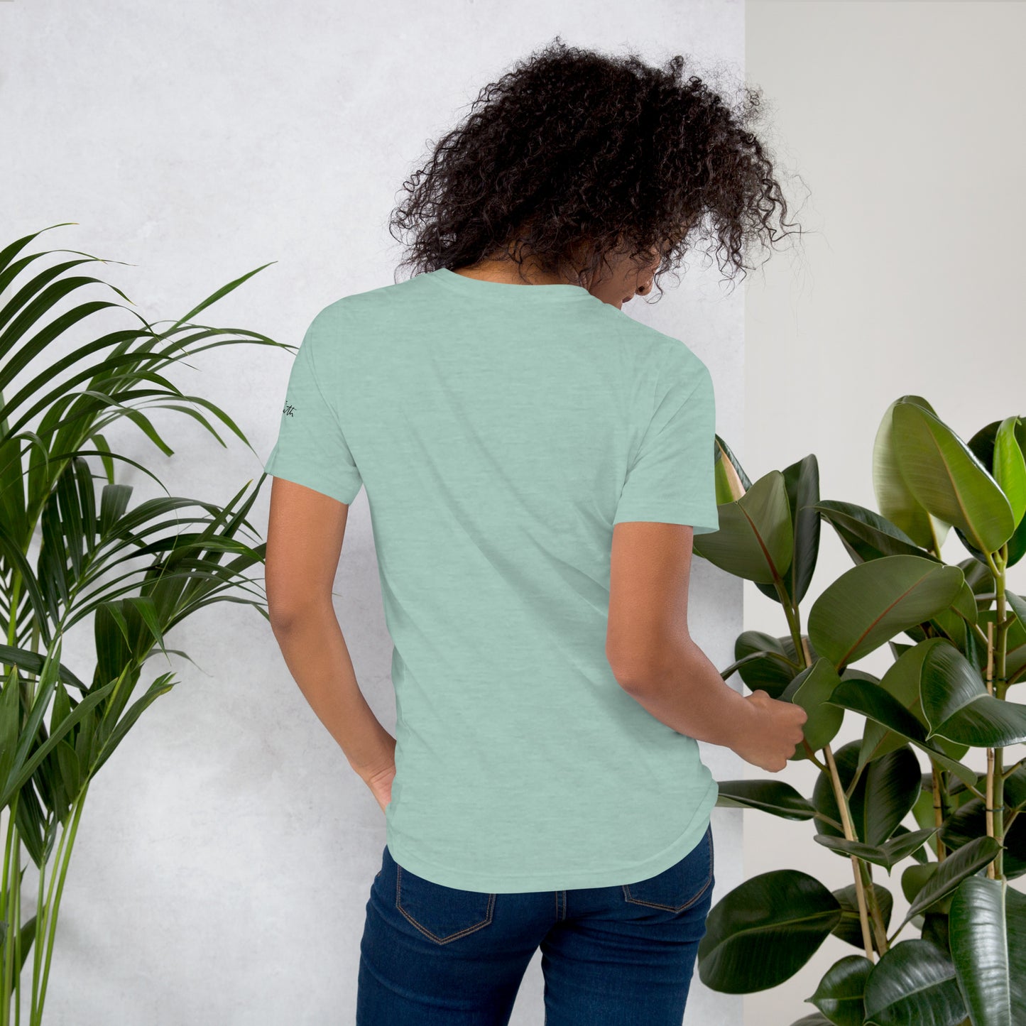 Person wearing a light green t-shirt standing between two potted plants against a white wall.