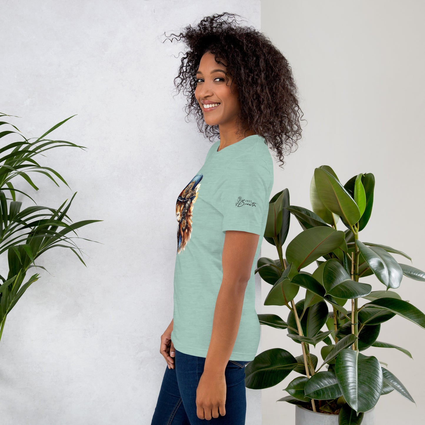 Woman wearing a light green t-shirt with a graphic design, standing between two potted plants against a white wall.