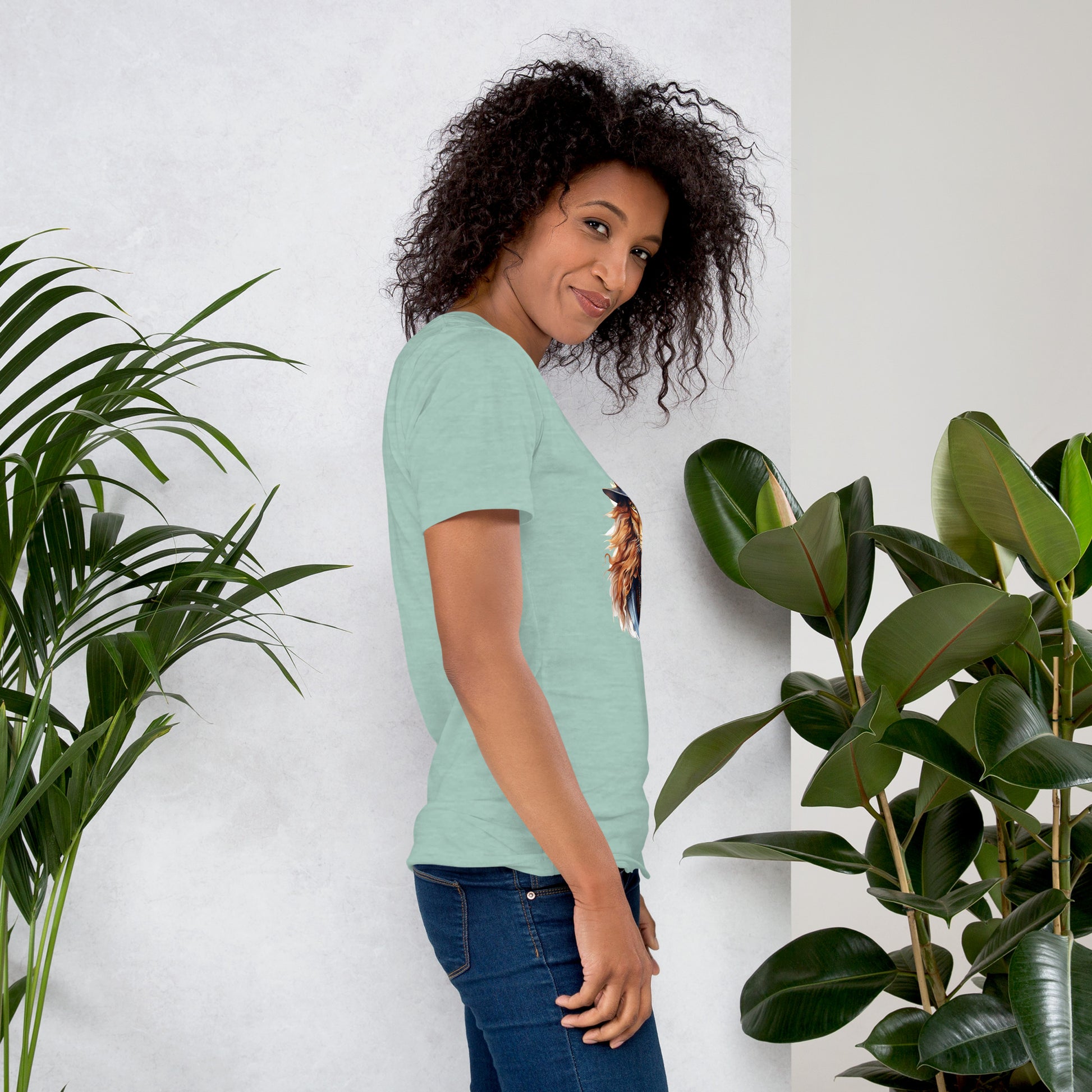 Woman wearing a green t-shirt with a graphic design, standing between two plants against a white wall.